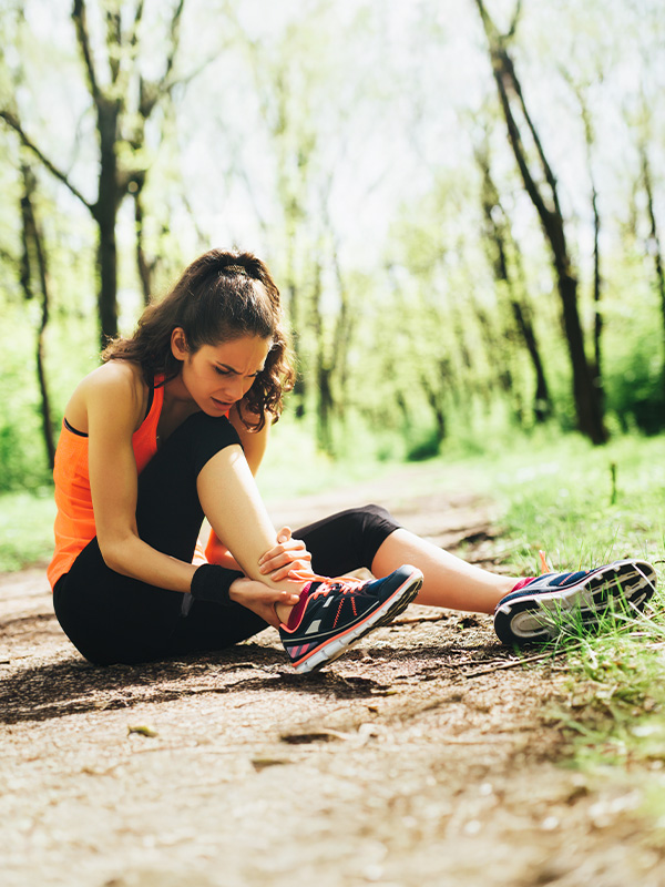 A runner sitting on the ground on a trail, grabbing her ankle in pain